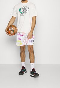 Helle, übergroße Herren-T-Shirt in Creme mit grafischem Druck, weiße Shorts mit Blumenmuster und schwarze Sportschuhe mit bunten Akzenten, während er einen Basketball hält.