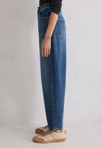 Personne portant un jean bleu droit taille haute et des baskets beiges avec des panneaux texturés, se tenant de côté contre un mur blanc uni.