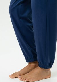 Marineblauwe joggingbroek met elastische boorden, gemaakt van zachte stof, met een gladde textuur en een ontspannen pasvorm, gedragen met blote voeten.