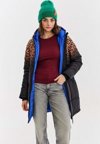 Žena v zelenej pletenej čiapke, čiernej páperovej bunde s leopardím vzorom a modrým podšívkou, vínovej košeli a svetlošedých džínoch, stojaca na bielom pozadí.