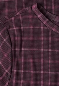 Gros plan d’un tissu à carreaux violet et noir montrant un col rond et une couture d’épaule sur un vêtement à texture douce.