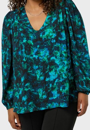 Frau trägt eine langärmelige V-Ausschnitt-Bluse mit abstraktem Muster in Blau, Grün und Schwarz, kombiniert mit einer schwarzen Hose.