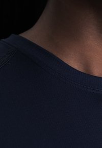 Κοντινό πλάνο στον λαιμό και τον ώμο με σκουρόχρωμο δέρμα που φοράει μπλε μπλούζα με στρογγυλή λαιμόκοψη σε σκουρόχρωμο φόντο.