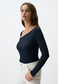 Body in jersey blu navy a maniche lunghe con scollo a barca e piccolo dettaglio metallico, abbinato a pantaloni bianchi a vita alta.