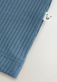 Ribbigt blått tyg med en mjuk textur. Har en liten vit etikett med ett smileyansikte. Fokus på kanten och sömmarnas detaljer.