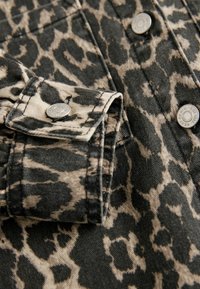 Látka s leopardím vzorom v hnedej a čiernej farbe s golierom a striebornými cvokmi. Detailný záber na štýlový manžetový rukáv z textúrovanej džínoviny.