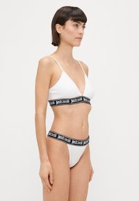 Bijeli bikini set koji se sastoji od trokutaste bralette i tangice, oba s crnim elastičnim trakama s natpisom "justcavalli". Glatka tekstura i minimalistički dizajn.