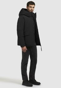 Sorte polstret jakke med stor hætte, frontlynlås og sidelommer; båret over mørke jeans og sorte sko. Glat tekstur.
