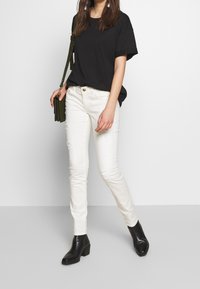Camiseta negra de gran tamaño, jeans skinny blancos desgastados y botines negros de tacón. También incluye un bolso cruzado verde.