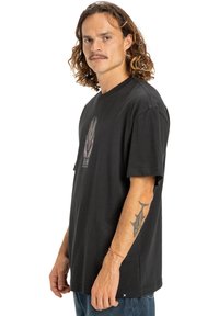 Čierne bavlnené tričko s krátkymi rukávmi, ktoré má farebný grafický dizajn na hrudi a uvoľnený strih. Modré džínsy, bočný pohľad.