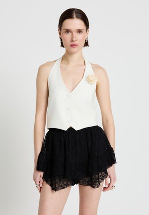 Donna che indossa un gilet halter bianco con bottoni e una spilla a forma di fiore abbinata a pantaloncini con gonna a strati in pizzo nero, in piedi davanti a uno sfondo bianco.