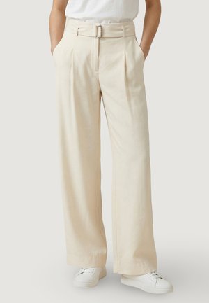 Persoon draagt crèmekleurige broek met hoge taille en wijde pijpen met bijpassende riem en witte sneakers, staand met handen in de zakken.