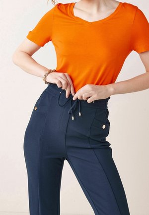 Personne portant un haut à manches courtes orange vif et un pantalon taille haute bleu marine, ajustant le cordon à la taille, avec un bracelet en or au poignet gauche.