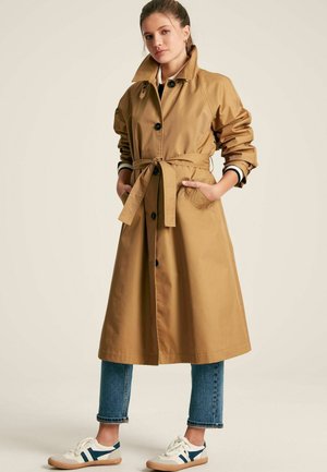 Khaki trench coat with a belt, large collar, and black buttons. Paired with cropped jeans and white sneakers featuring blue accents.