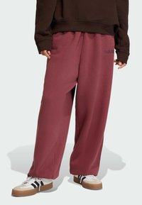 Pantalones de chándal anchos color burdeos hechos de una tela suave, con una cinturilla elástica y un pequeño logo negro de adidas en el lateral.
