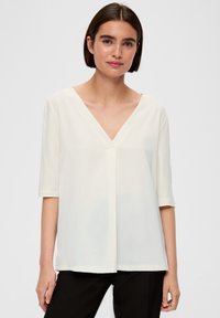 Biała bluzka z krótkim rękawem o dekolcie w kształcie litery V, luźny krój i gładka tekstura; posiada delikatne plisowanie na środku i prosty dół.