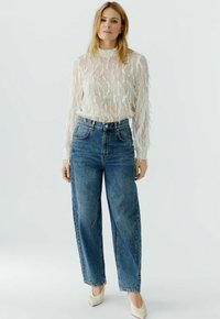 Sheer white long-sleeve top with textured fringe detail, paired with high-waisted blue denim wide-leg jeans and nude heels.