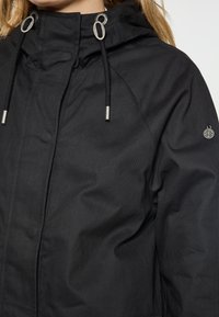 Giacca leggera nera con cappuccio, dettagli argentati sul cordino e logo sulla manica. Tessuto liscio con chiusura a zip davanti.