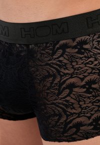 Czarne koronkowe majtki typu boxer z solidnym czarnym elastycznym paskiem w talii z wytłoczonym logo "HOM". Materiał ma kwiatowy wzór i półprzezroczystą teksturę.