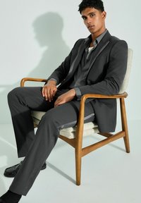 Costume gris au design ajusté, avec un revers en satin. Le modèle est assis sur une chaise en bois, le pantalon et la veste sont ajustés.