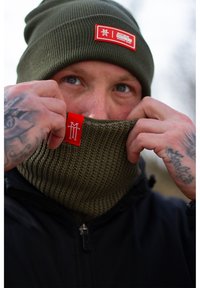 Green knit beanie and matching neck gaiter, both with red logo tags. The texture is ribbed, and the fabric appears warm and flexible.