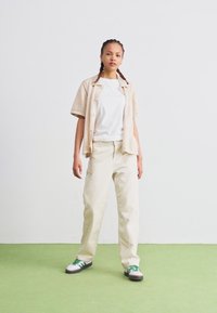 Beige short-sleeve shirt over a white t-shirt, paired with light-colored pants and green-accented sneakers, on a green floor.