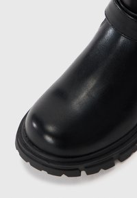 Botte cheville en cuir noir avec un bout arrondi, une surface texturée et une semelle en caoutchouc robuste dotée de motifs prononcés.