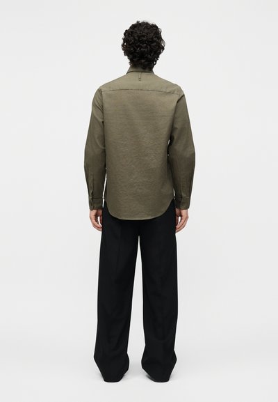 Personne aux cheveux bouclés portant une chemise à manches longues vert olive et un pantalon noir ample, debout dos à un fond uni.