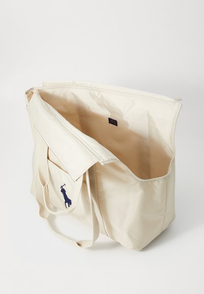 Beige kanvas tote soma ar garām siksnām, atvērta augšpuse, ar iekšējo birku un tumši zilas žirafes grafiku sānos.
