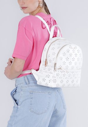 Mujer con pendientes de aro dorados, camisa rosa, vaqueros azul claro y mochila blanca con estampado floral y cremalleras doradas en la espalda.