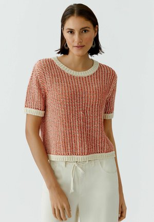 Frau trägt roten und weißen Kurzarm-Strickpullover mit cremefarbenen Kanten und cremefarbenen Jogginghosen mit Kordelzug, steht vor einfarbigem Hintergrund.