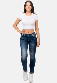 Jeans attillati di colore blu scuro con una leggera sbiaditura, cinque tasche e chiusura con bottone, abbinati a una t-shirt bianca corta e scarpe da ginnastica bianche.