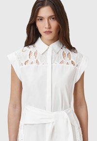 Weißes Hemdkleid mit Spitzeneinsätzen an den Schultern und der Brust, kurzen Ärmeln und einem Bindeband an der Taille. Glattes Material, taillierte Gestaltung.