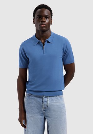 Mann trägt ein blaues kurzärmeliges Poloshirt mit Frontreißverschluss, kombiniert mit hellblauen Jeans, steht vor einem einfarbigen Hintergrund.