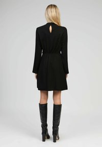 Femme avec de longs cheveux blonds portant une robe noire à manches longues et des bottes noires à talons hauts jusqu'aux genoux, se tenant de dos sur un fond uni.
