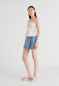 Top bianco in pizzo senza spalline con orlo a strati, abbinato a pantaloni shorts in denim azzurro chiaro. La modella indossa sandali a scivolo rosa chiaro, in piedi davanti a uno sfondo neutro.