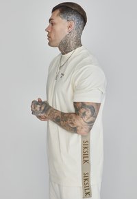 T-shirt di colore crema con maniche corte, caratterizzata da un logo sottile e da una striscia beige con il marchio lungo il lato. Tessuto morbido, vestibilità rilassata.