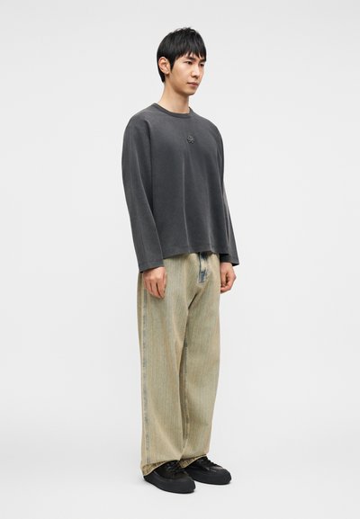 Σκούρο γκρι μακρυμάνικη μπλούζα με στρογγυλή λαιμόκοψη, συνδυασμένη με τζιν φαρδιά παντελόνια σε ανοιχτό μπεζ χρώμα με κάθετες ρίγες. Μαύρα παπούτσια ολοκληρώνουν την εμφάνιση.