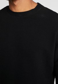 Maglione nero in maglia con un design a coste testurizzato e colletto tondo, che mostra un primo piano dei dettagli del tessuto e del collo.