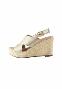 Sandale compensée en espadrille à lanières croisées dorées avec un talon en jute texturé et une fermeture à boucle. Conception à bout ouvert avec un dessus lisse.