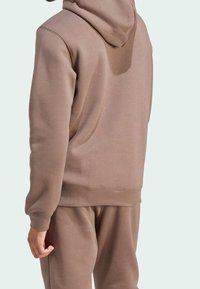 Osoba v hnědé mikině a teplácích, zobrazená zezadu na jednobarevném světlém pozadí.