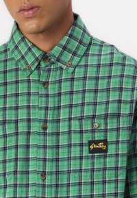 Green plaid button-up shirt made of woven fabric, featuring brown buttons and a black label with yellow text on the left chest pocket.