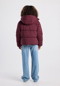 Bordeaux pufferjack met capuchon, voorzien van gestikte secties, gecombineerd met lichtblauwe wijde jeans in een casual houding.