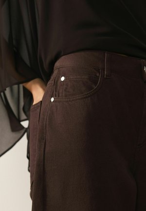 Woman wearing dark brown jeans with metal rivets, hand partially in pocket, and a sheer black top with flowing sleeves.