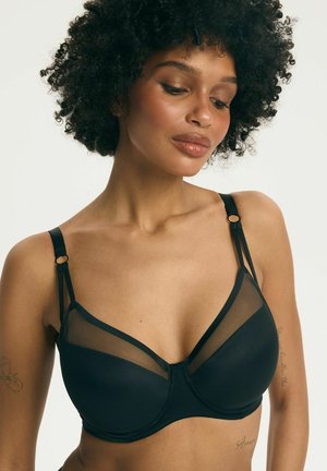 Kobieta o kręconych włosach, ubrana w czarny biustonosz z przezroczystymi panelami z siateczki, patrząca w dół, na jasnym, jednolitym tle.