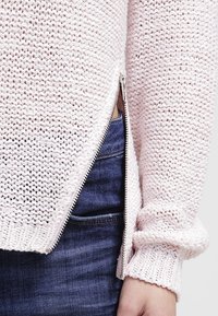 Pullover in maglia rosa chiaro con trama testurizzata, dettaglio della zip laterale e polsini a costine, abbinato a jeans blu.
