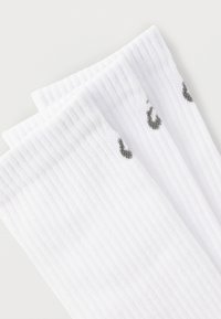 Τρία ζευγάρια λευκές ριγωτές κάλτσες μεσαίου μήκους, τακτοποιημένα στοιβαγμένα, με μικρά γκρι λογότυπα κοντά στην κορυφή, σε λευκό φόντο.