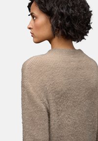 Nainen, jolla on lyhyet kiharat tummat hiukset, pukeutuneena vaaleanbeigeen neulepaitaan, kuvattuna selkä vasemmalle käännettynä neutraalilla taustalla.