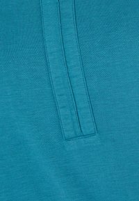 Tela de camisa color azul verdoso con un acabado texturizado, que presenta un detalle de costura vertical. El material parece suave y elástico, con un tono suave y uniforme.