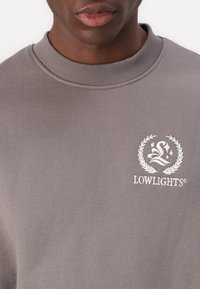 Grå hettegenser med ribbestrikket halslinning og en hvit brodert logo med stilisert design og laurbæraksenter på brystet.